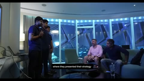 Source frame at 155.500s. Interview, student laptop groups, and glass-walled presentation room.